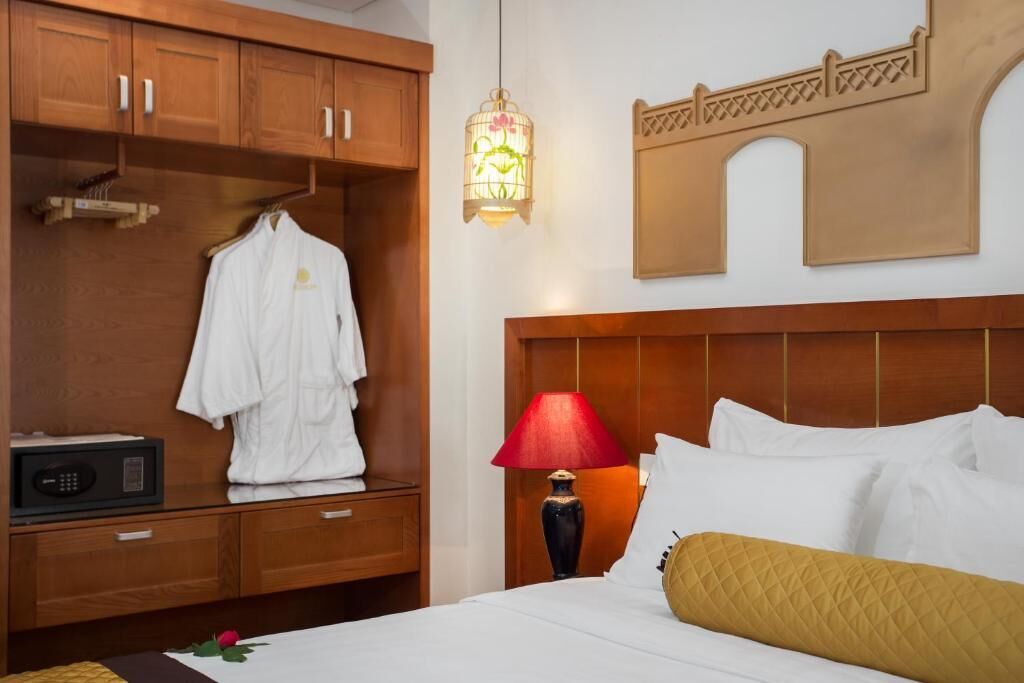 A hotel room with a wooden wardrobe, a bed with white bedding and a yellow pillow, a red lamp, and a decorative wall piece.