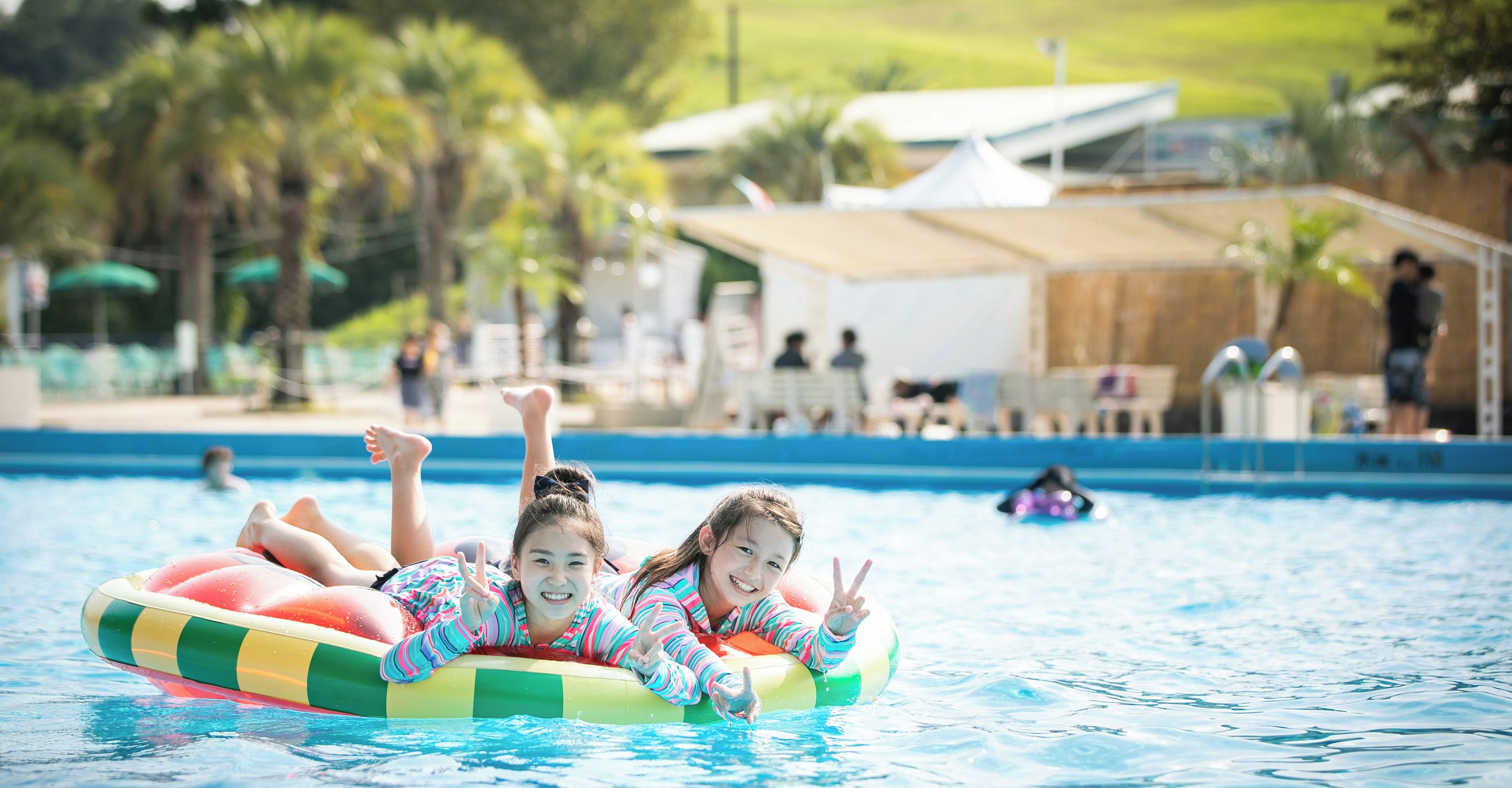 Two individuals floating on an inflatable pool float in a swimming pool with a clear view of the surrounding area, including a building and trees.