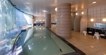 Un área de piscina interior moderna con una barrera de vidrio, suelo de baldosas y una fila de taburetes blancos.