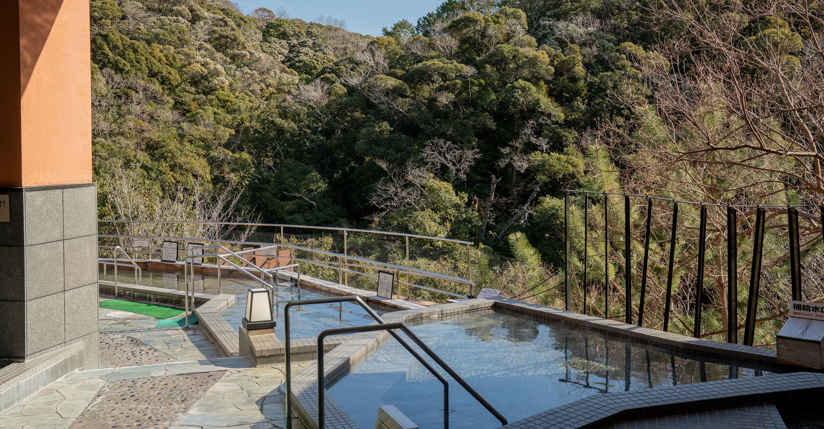 A pool area with a stone deck, metal railings, and a view of a forested hillside.