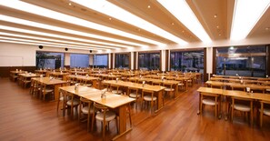 Un amplio salón de conferencias bien iluminado, con numerosas mesas y sillas, techos altos y grandes ventanales.