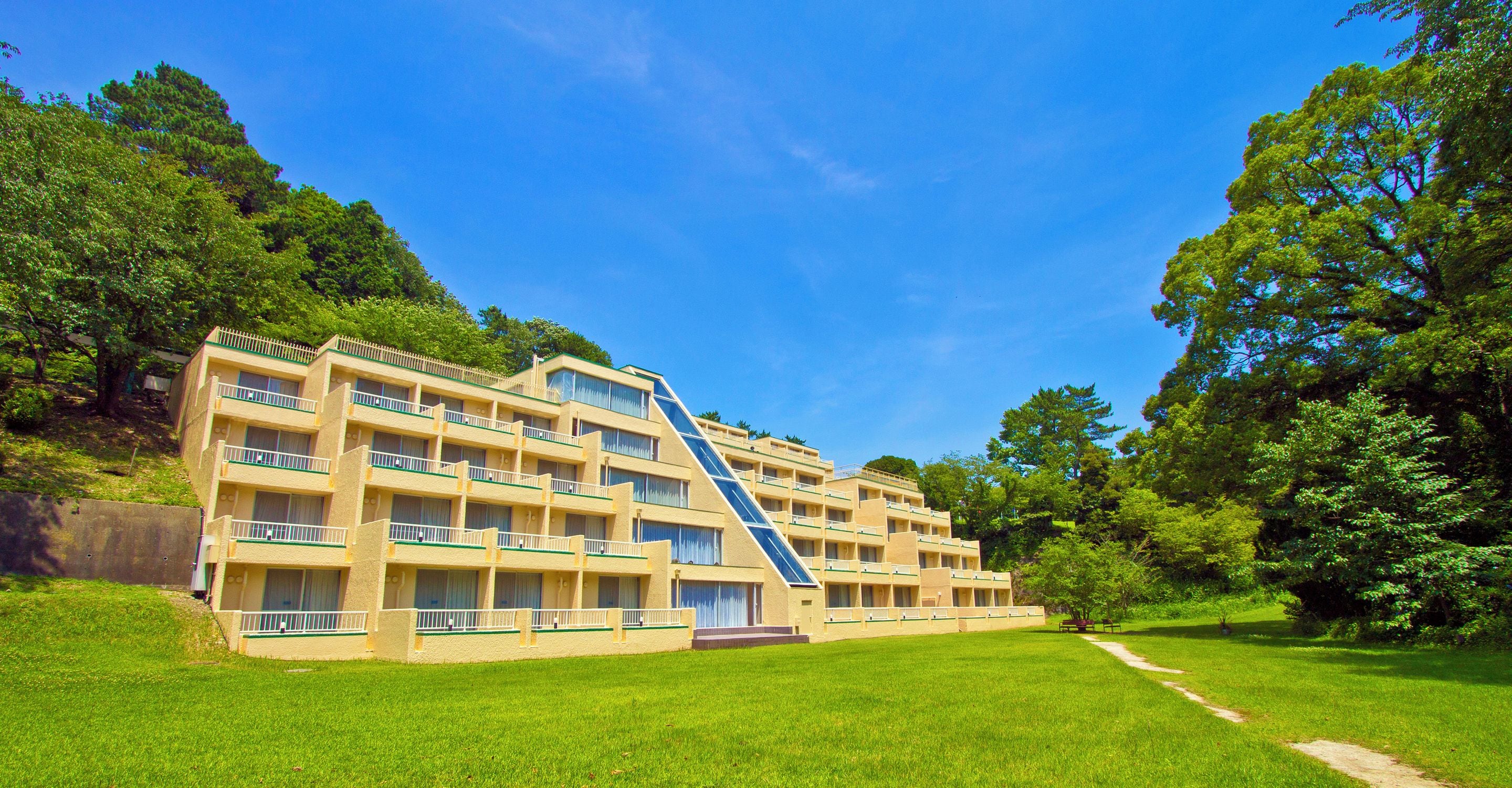 A multi-story hotel with a central staircase, surrounded by lush greenery and a clear blue sky.