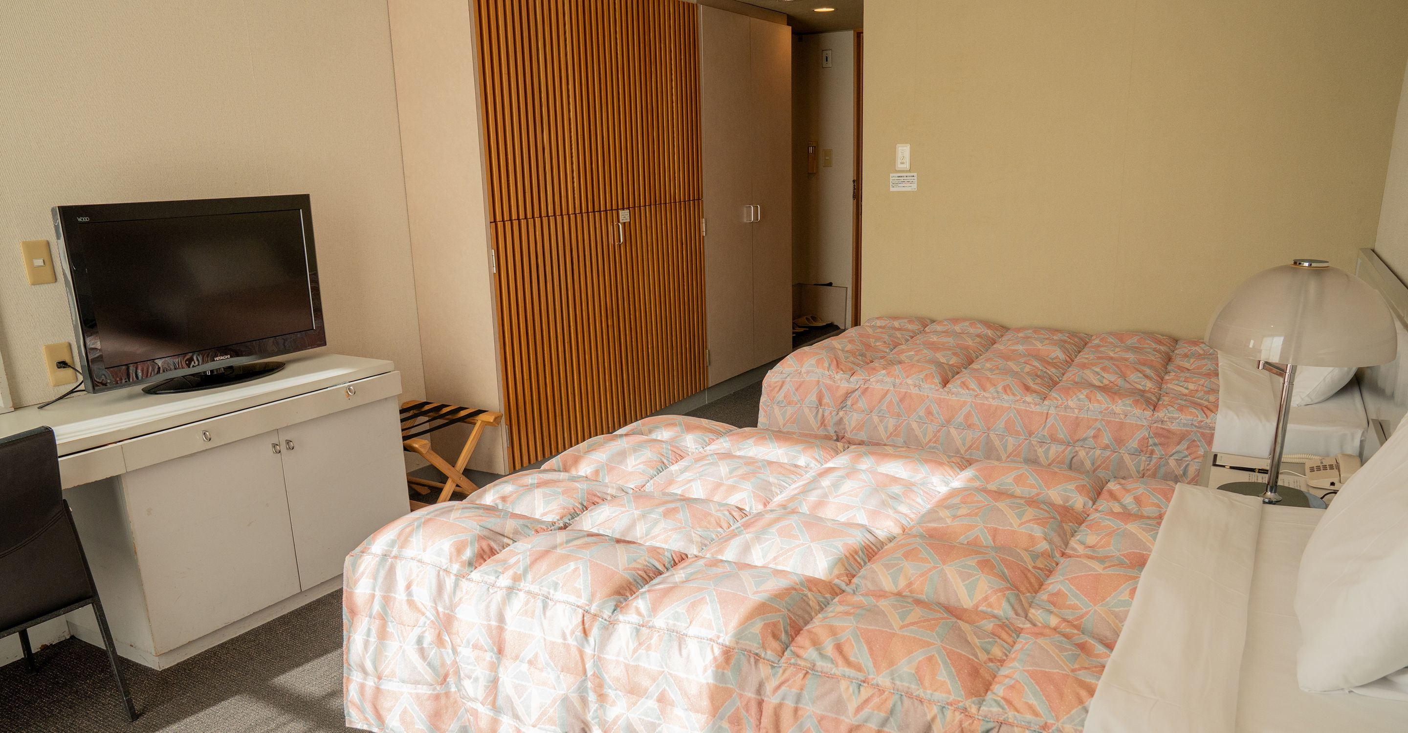 A hotel room with a television on a desk, a bed with patterned bedding, a bedside lamp, and a wardrobe.
