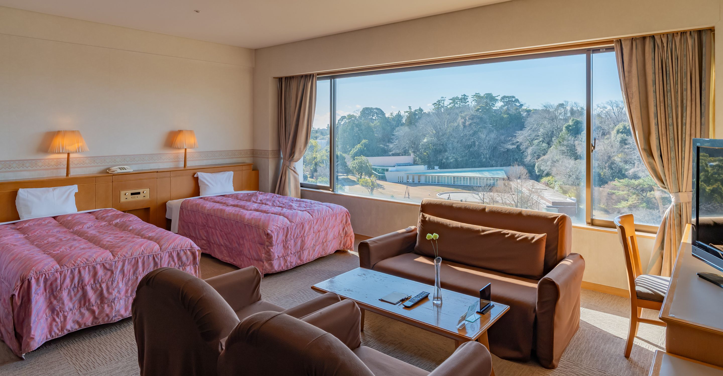 A hotel room with two beds, a sofa, a coffee table, a TV, and a view of a building and trees.