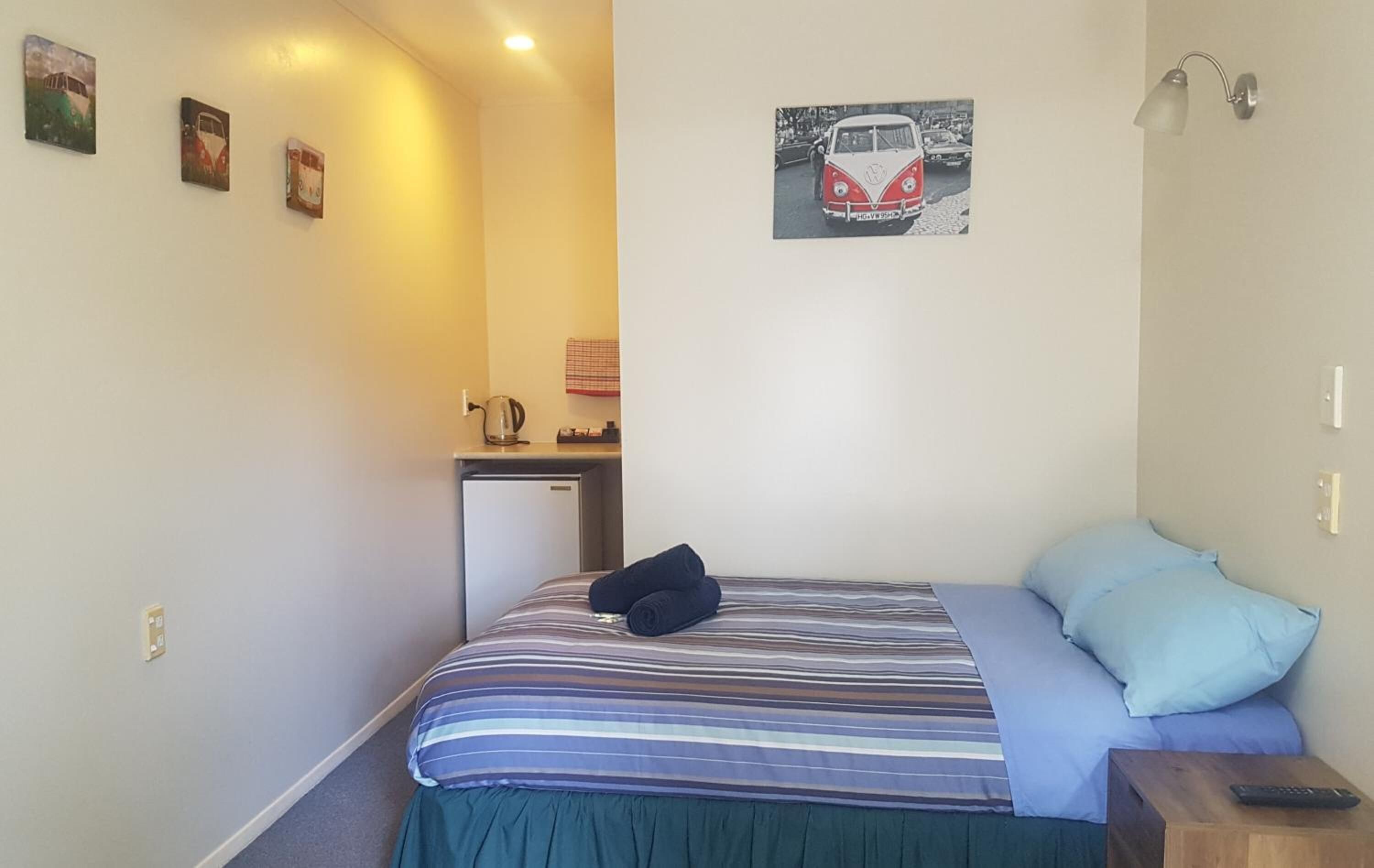 A single bed with a striped bedspread and a blue pillow, a small bedside table with a remote control, and a kitchenette area with a kettle and a small refrigerator.