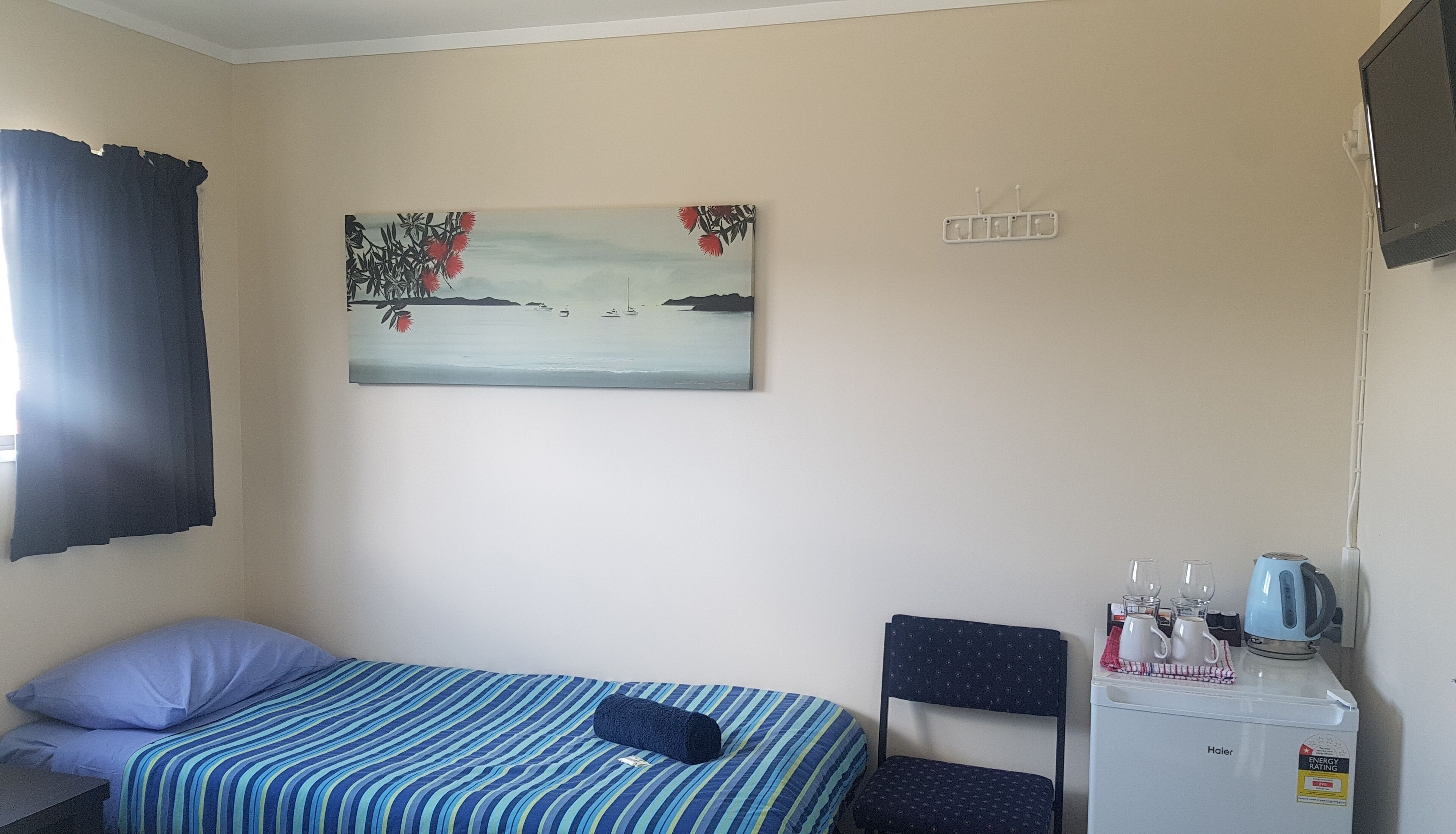 A bedroom with a bed, a chair, a small table with a kettle, a coffee maker, and a painting on the wall.
