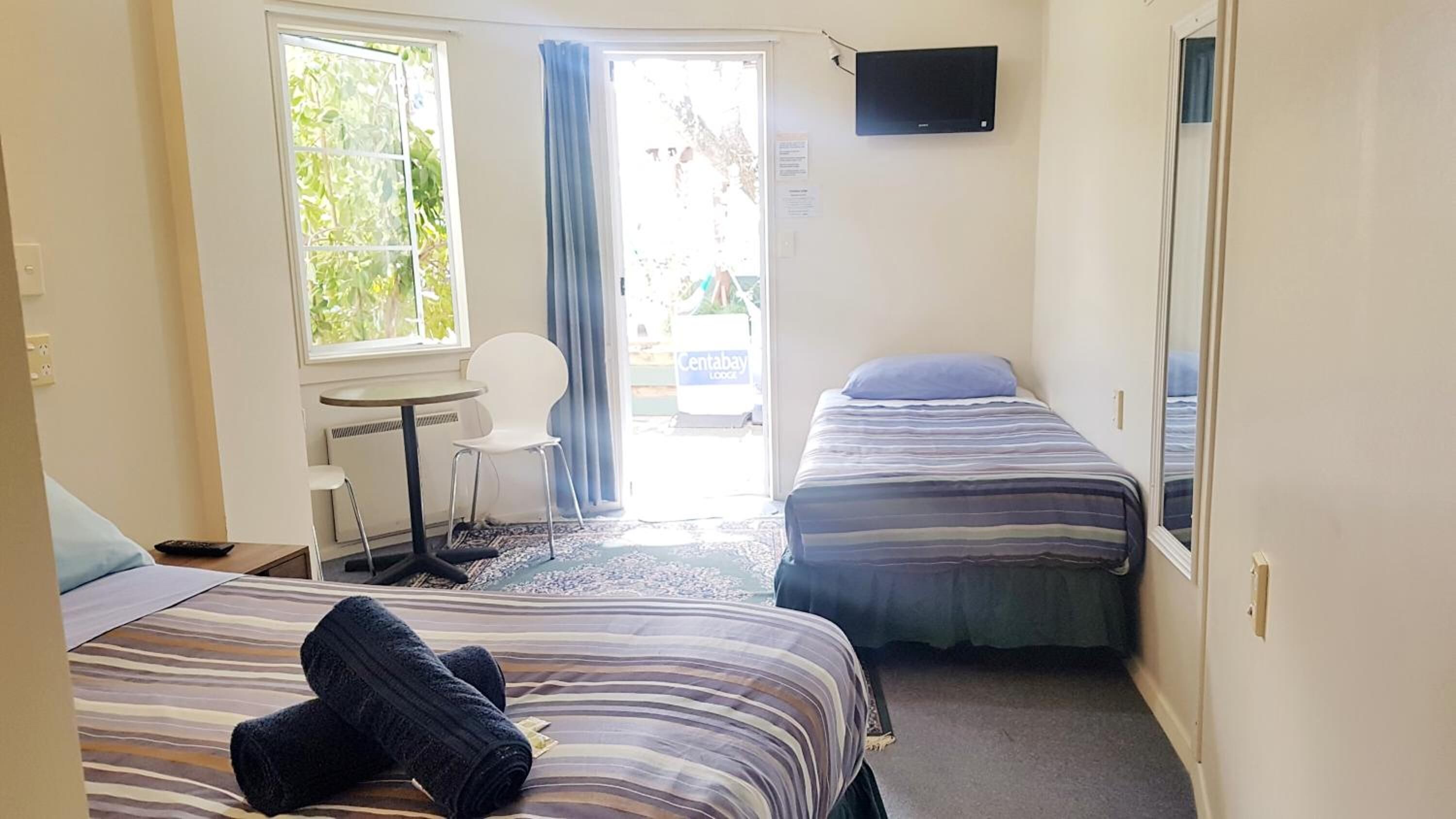 Une chambre d’hôtel avec deux lits, une petite table avec une chaise, un téléviseur fixé au mur et une fenêtre avec des rideaux.