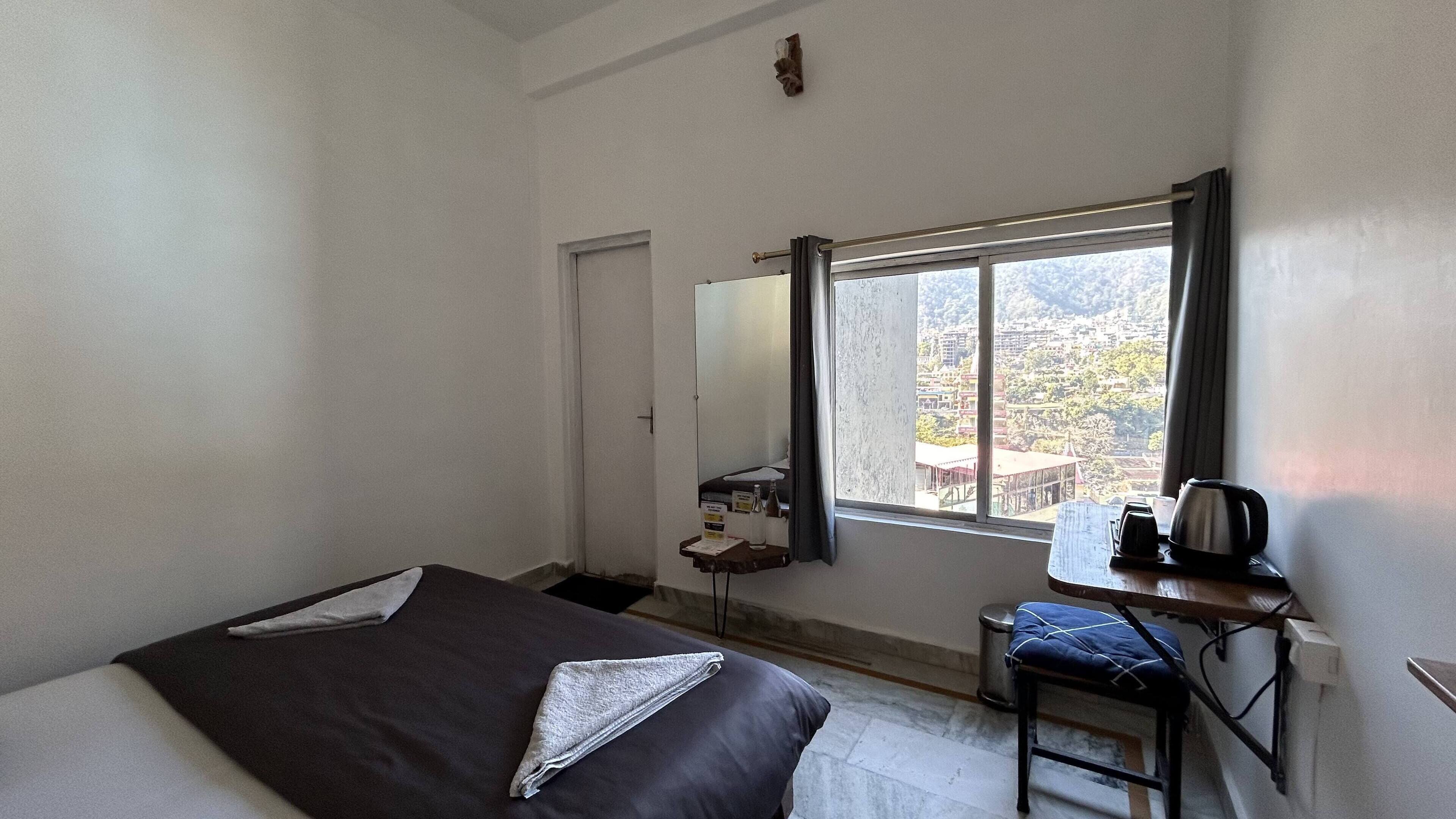 A small, sparsely furnished room with a bed, a desk, and a window offering a view of a mountainous landscape.
