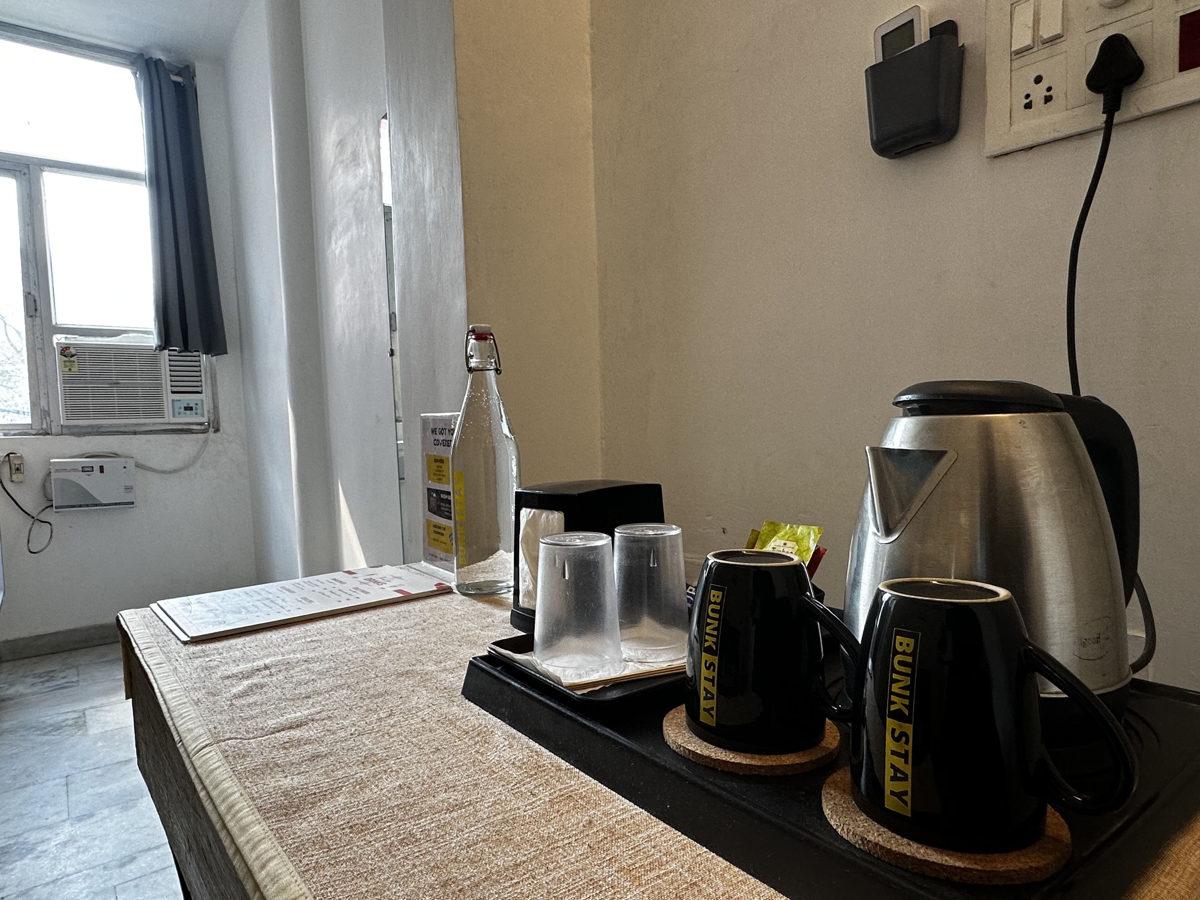 A hotel room with a bed, a coffee maker, two mugs, a bottle, and a glass on a tray.