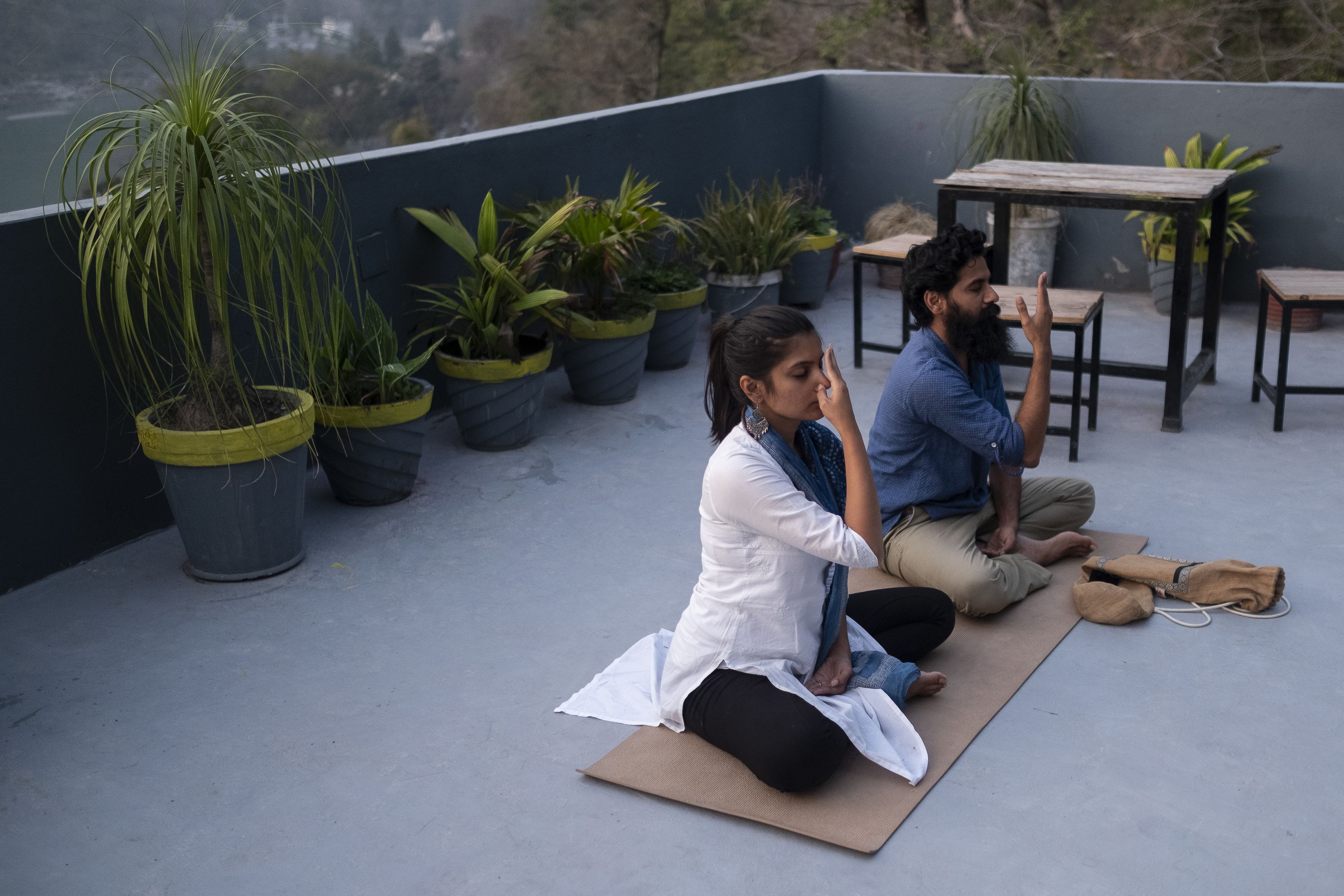Two people sitting on a yoga mat on a rooftop with potted plants around them.