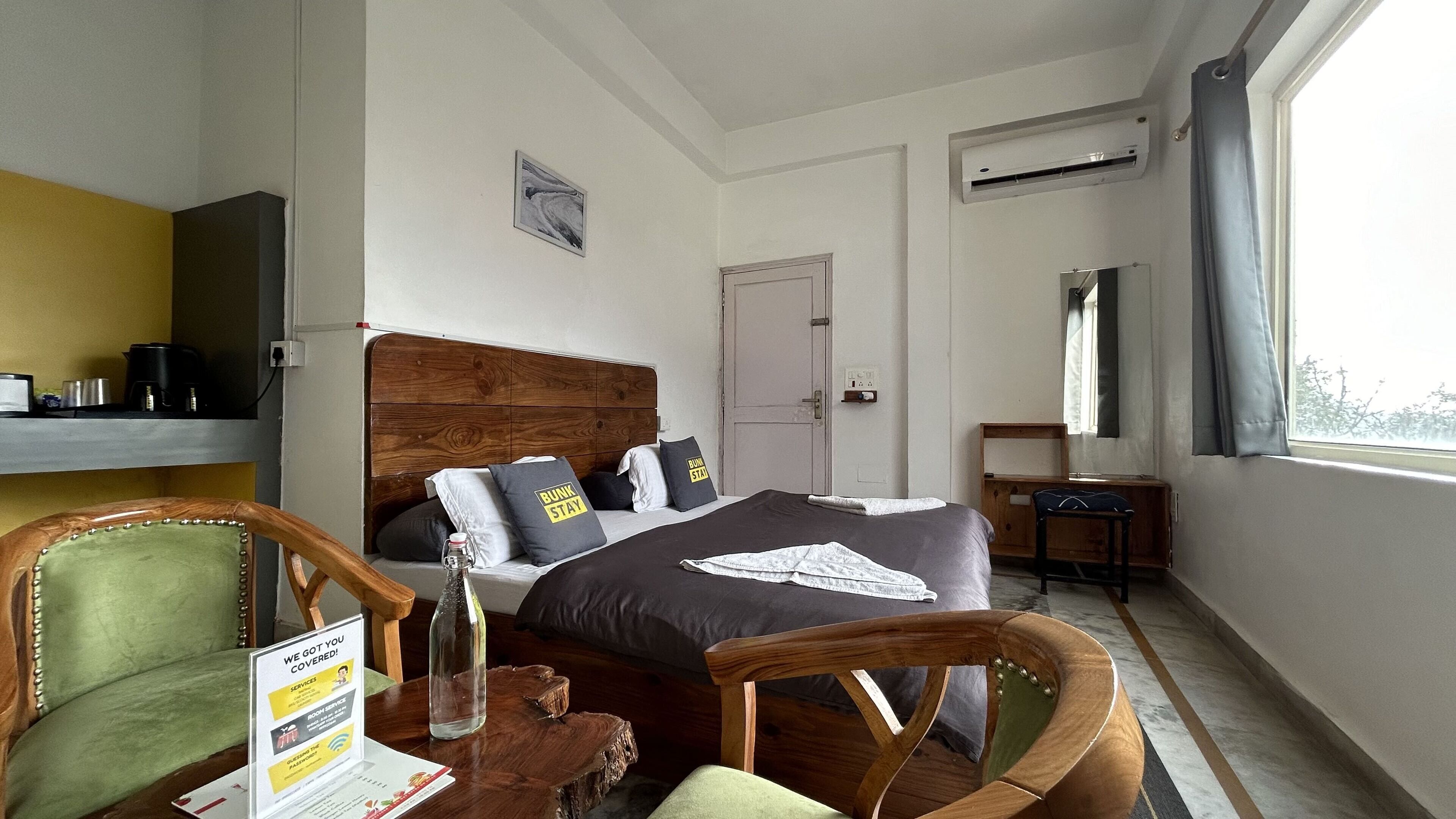 Une chambre d’hôtel avec un lit en bois, un bureau, une chaise, une petite table et une fenêtre avec des rideaux.