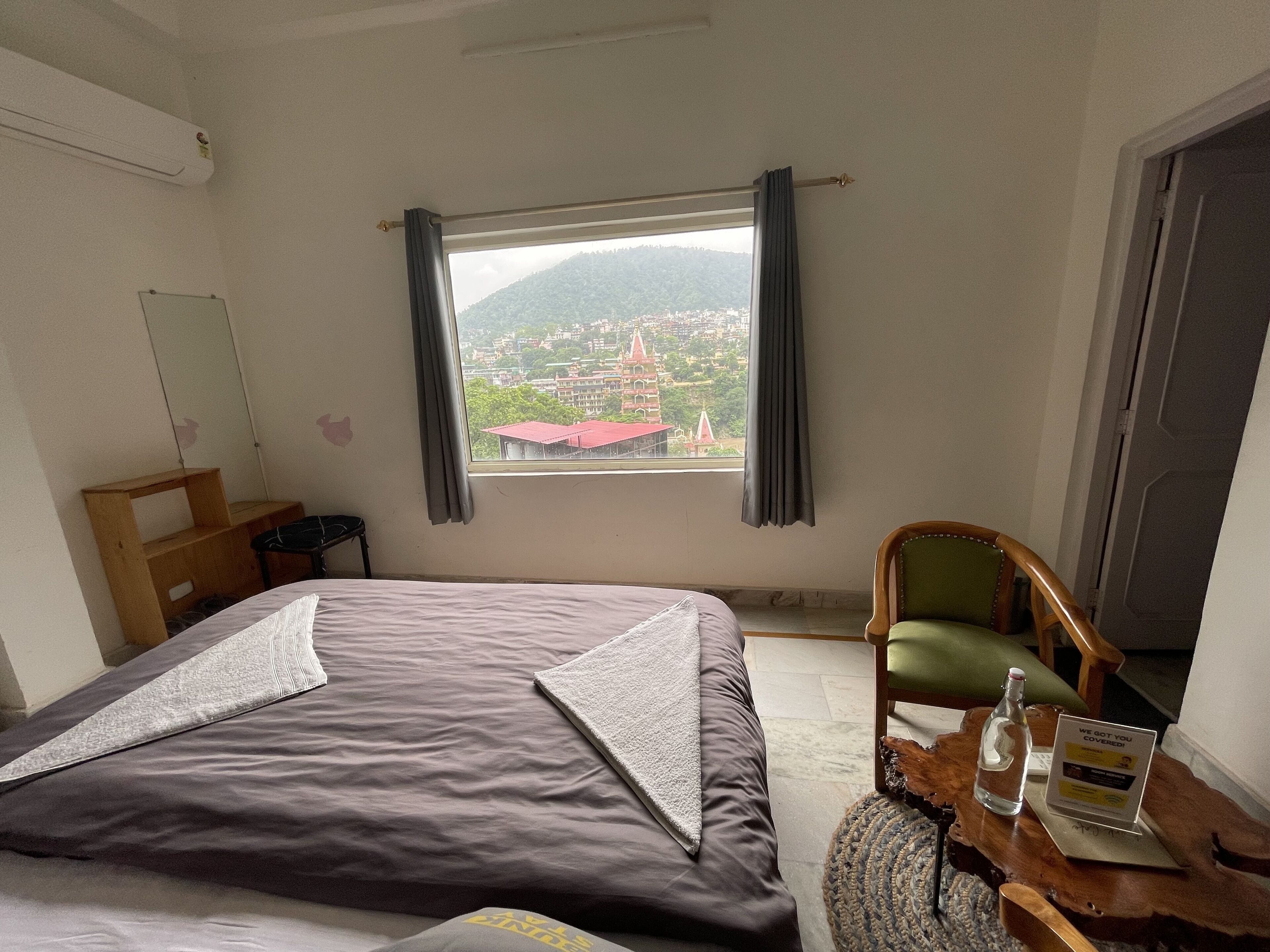 A hotel room with a bed, a wooden nightstand, a chair, a table with a bottle and a sign, and a window with a view of buildings and mountains.