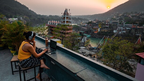 A person sitting on a balcony with a laptop, overlooking a town with buildings and a hill in the background. - Riverside home by bunkstay (Yamkeshwar)