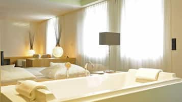 Une chambre d’hôtel avec une grande baignoire, une table de chevet avec une lampe et une fenêtre avec des rideaux légers.