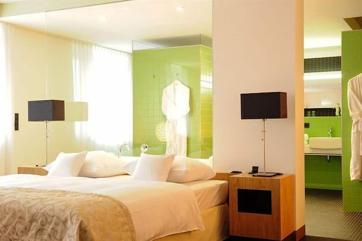 Une chambre d’hôtel avec un lit, des tables de chevet, un miroir, un peignoir accroché au mur et une salle de bain avec un lavabo et un miroir.