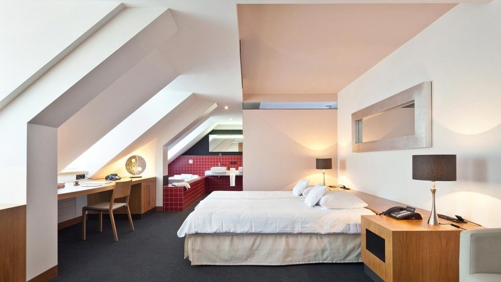Una habitación de hotel moderna con espacio de trabajo en altura, una cama con ropa blanca, un escritorio con silla y una mesita de noche con lámpara.
