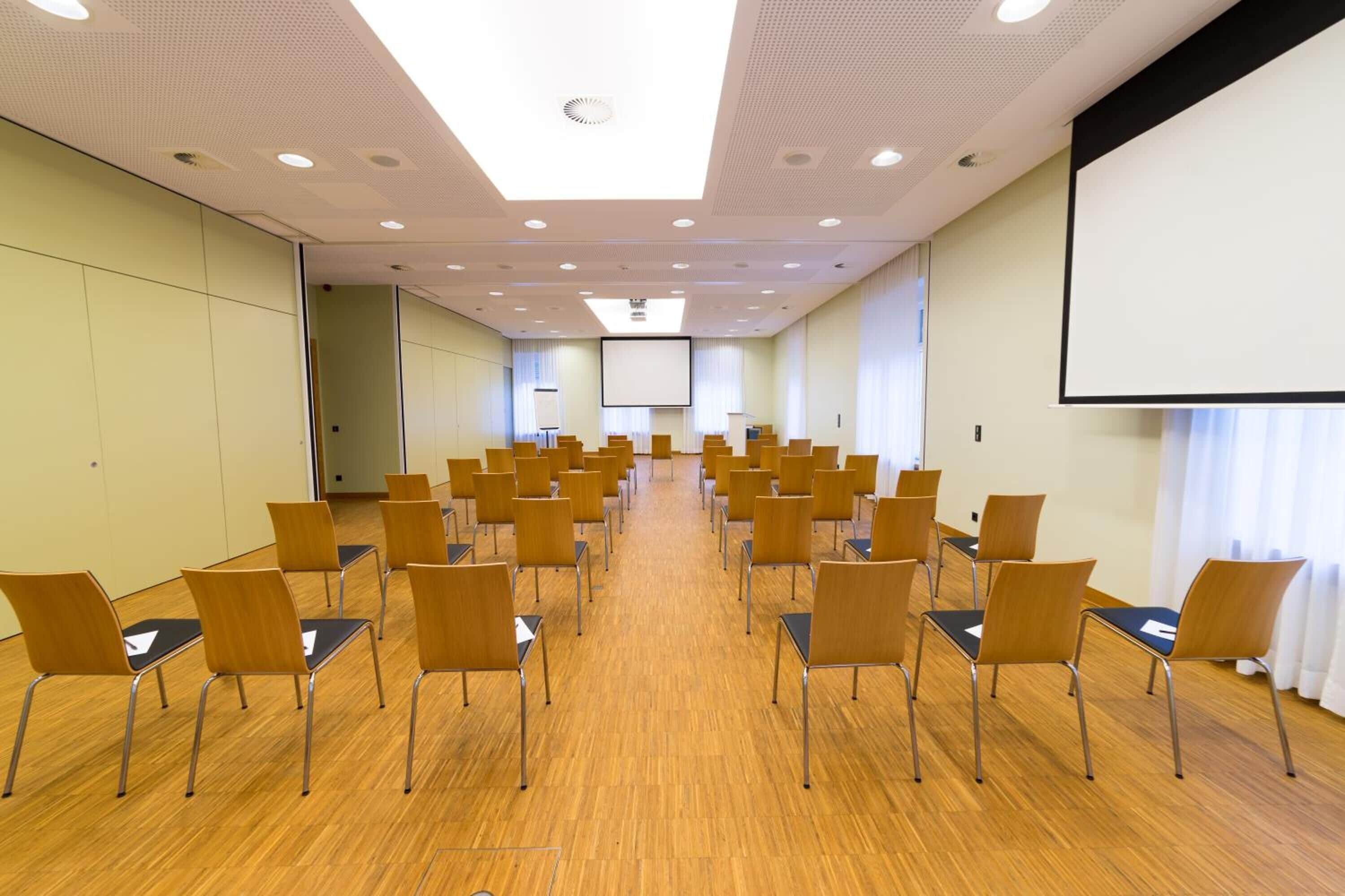 Un salón de conferencias con filas de sillas, una pantalla de proyector y una pantalla grande en la pared.
