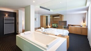 Habitación de hotel moderna con una bañera grande, una cama con ropa blanca, un televisor y un escritorio con lámpara.