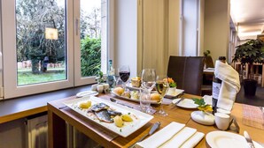 Mesa de comedor dispuesta con platos, vasos y una botella de vino, con vista a la vegetación exterior.