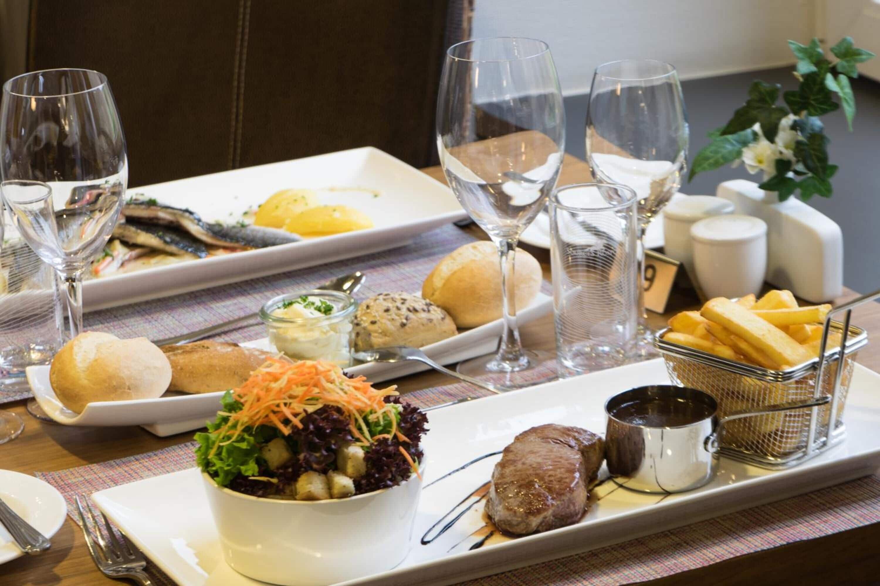 Een eettafel met diverse gerechten, waaronder vis, brood, salade en vlees, plus verschillende wijnglazen en een kleine schaal friet.