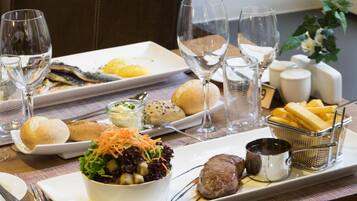 Een eettafel met diverse gerechten, waaronder vis, brood, salade en vlees, plus verschillende wijnglazen en een kleine schaal friet.