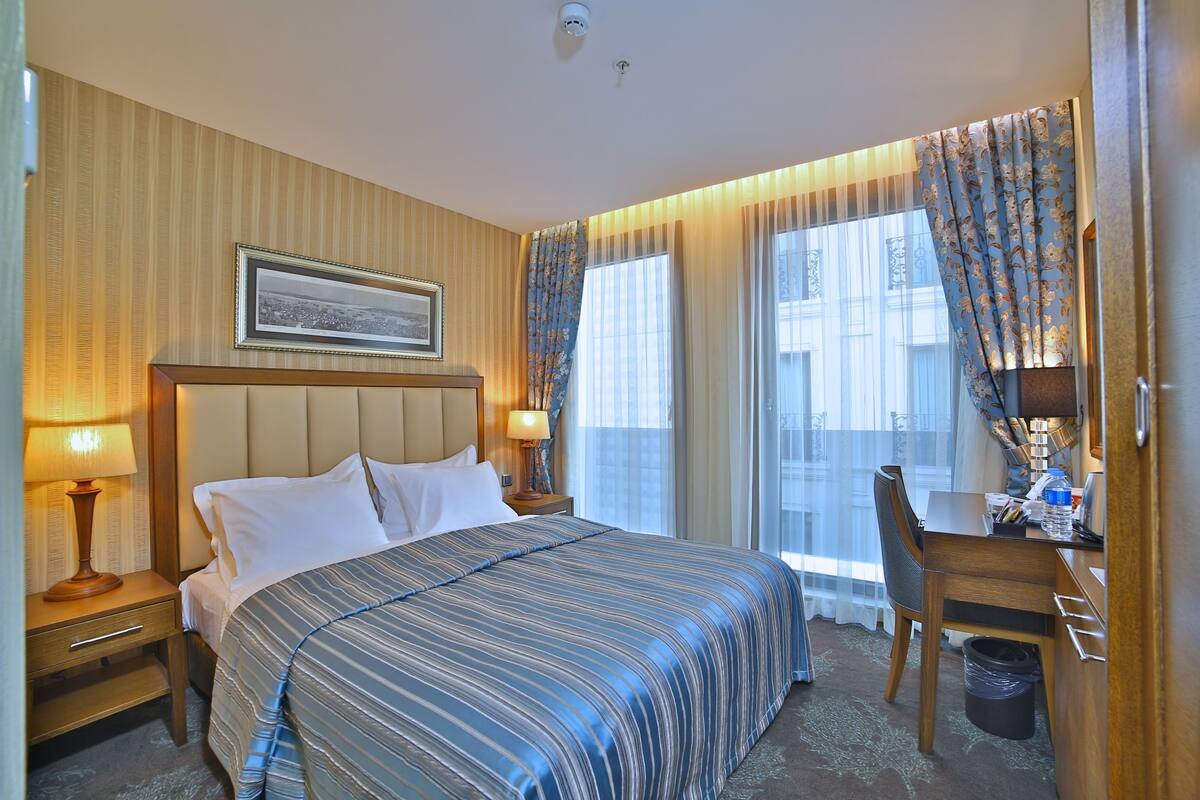 Une chambre d’hôtel avec un grand lit, un bureau et une chaise. Il y a un tableau au mur, une lampe sur la table de chevet et une fenêtre avec des rideaux.