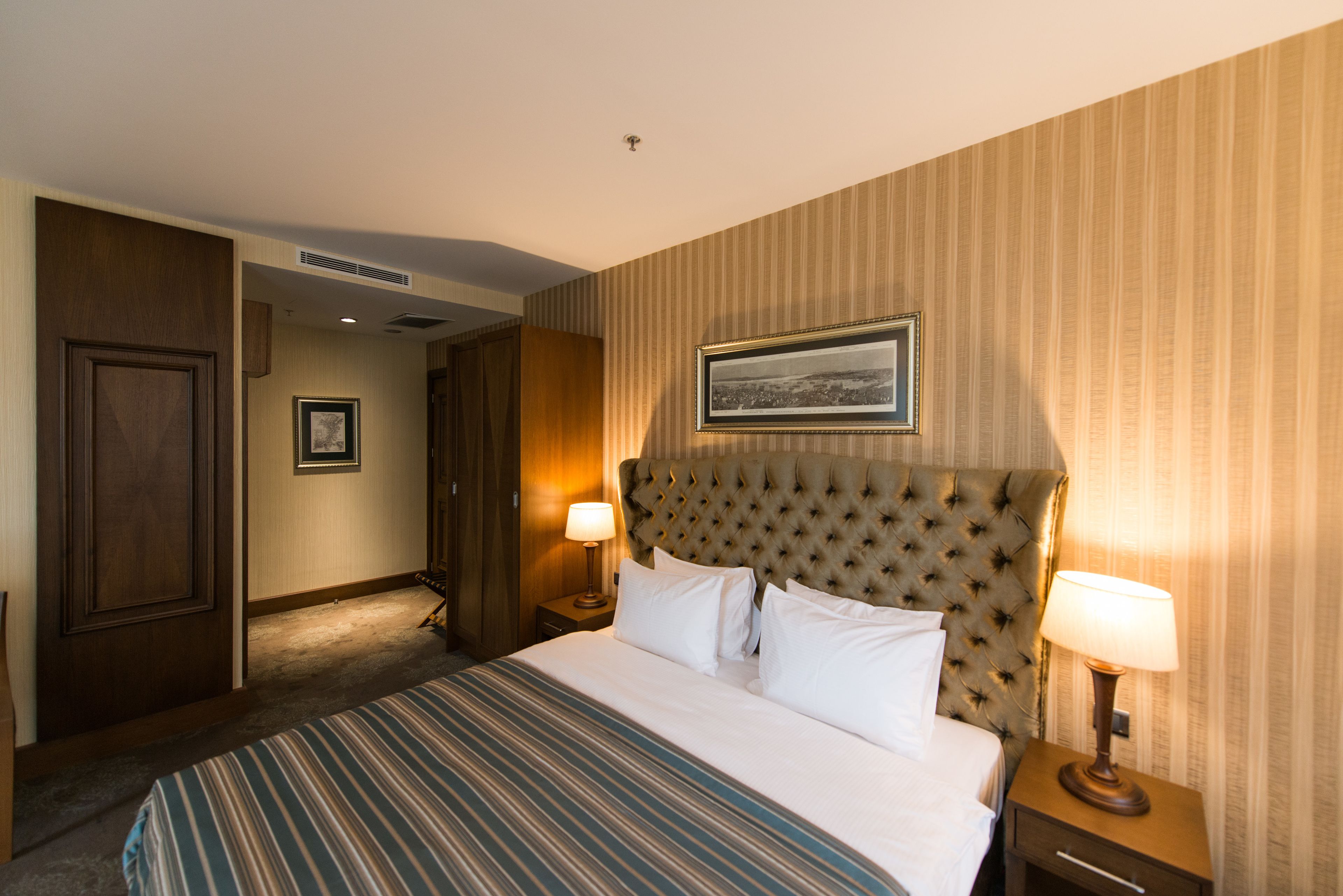 A hotel room with a large bed, a headboard, two bedside tables with lamps, a framed picture on the wall, and a door leading to another room.