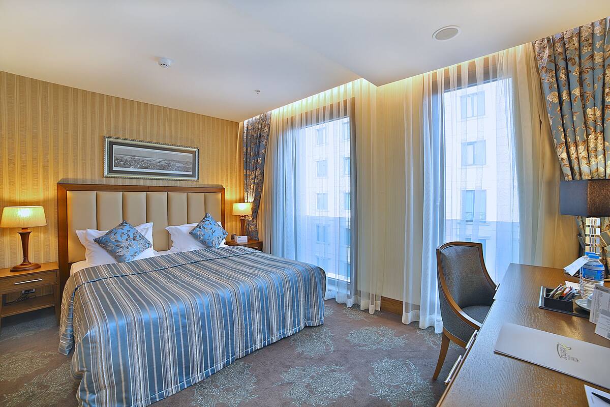 Une chambre d’hôtel avec un grand lit, un bureau et une chaise. Il y a une fenêtre avec des rideaux, un tableau au mur et une lampe sur la table de chevet.