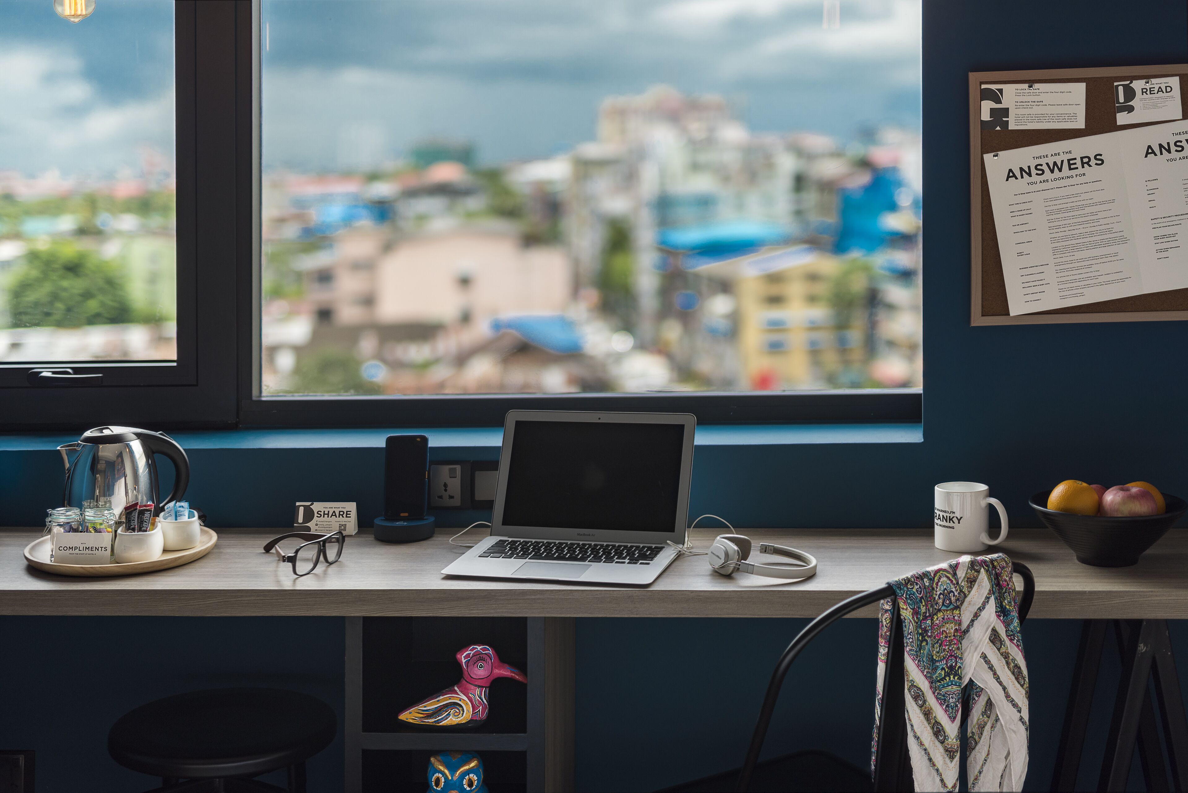 A desk with a laptop, a mug, a kettle, and a bowl of fruit. There is a window with a view of buildings and a bulletin board with a list titled "ANSWERS".
