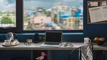 A desk with a laptop, a mug, a kettle, and a bowl of fruit. There is a window with a view of buildings and a bulletin board with a list titled "ANSWERS".