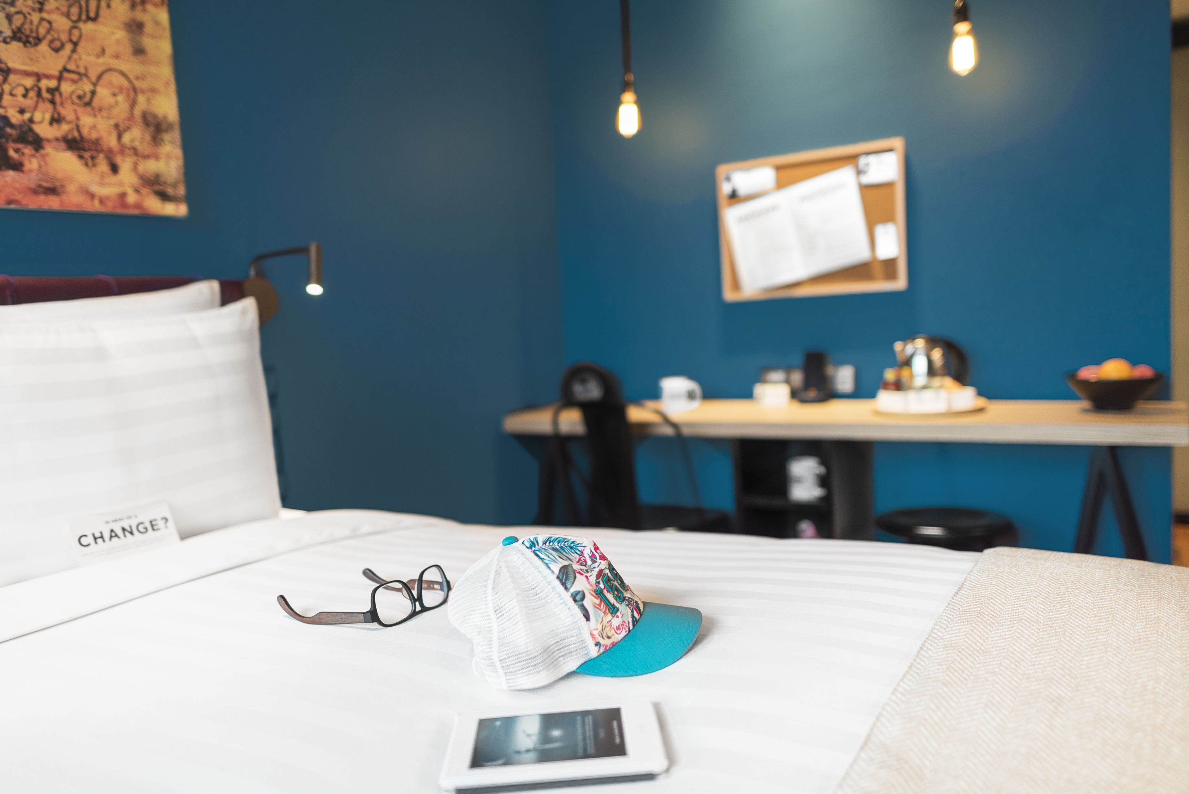 A neatly made bed with a pillow labeled "CHANGE?" and a cap on it. A tablet and glasses are also on the bed.