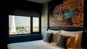 Ein Hotelzimmer mit großem Fenster und Blick auf die Stadt, einem Bett mit Kissen sowie einer Wand mit einem Wandgemälde mit dem Titel "The Liberation of Art".