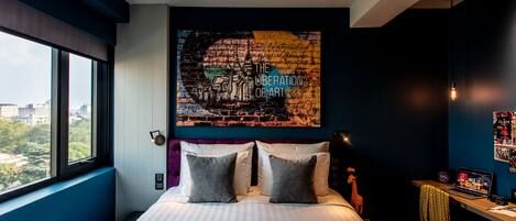 A modern hotel room with a large bed, a city view, a wall art piece titled "The Liberation of Art", and a desk with a laptop and a camera.