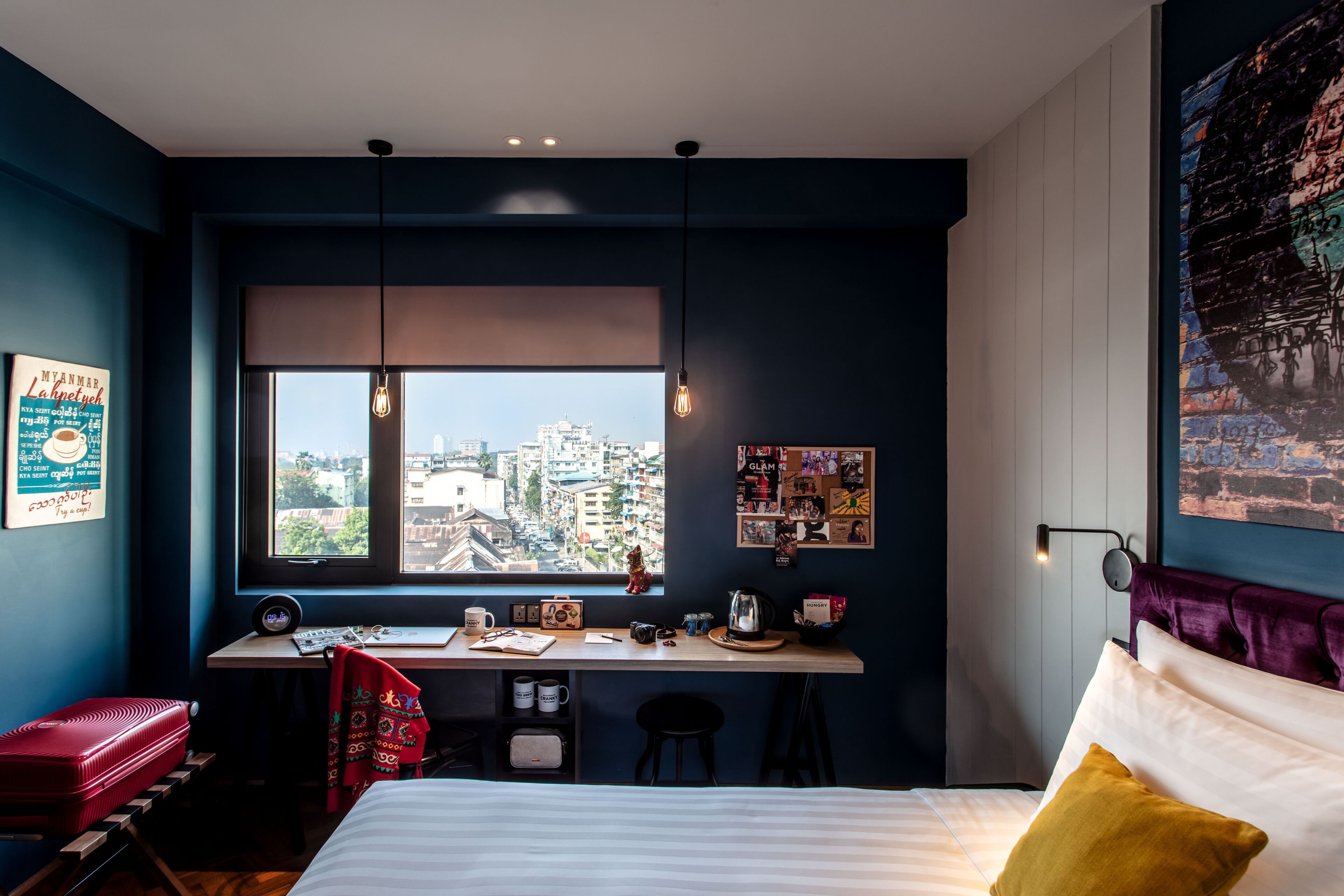 A bedroom with a city view, a desk with a chair, a bed with pillows, and a red suitcase.