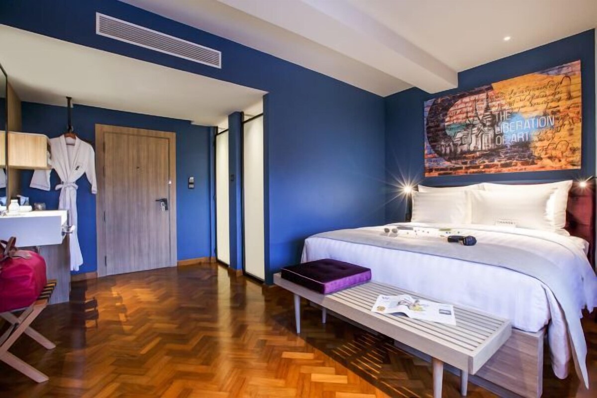 A hotel room with a bed, a bench, a table, a robe, and a painting on the wall.