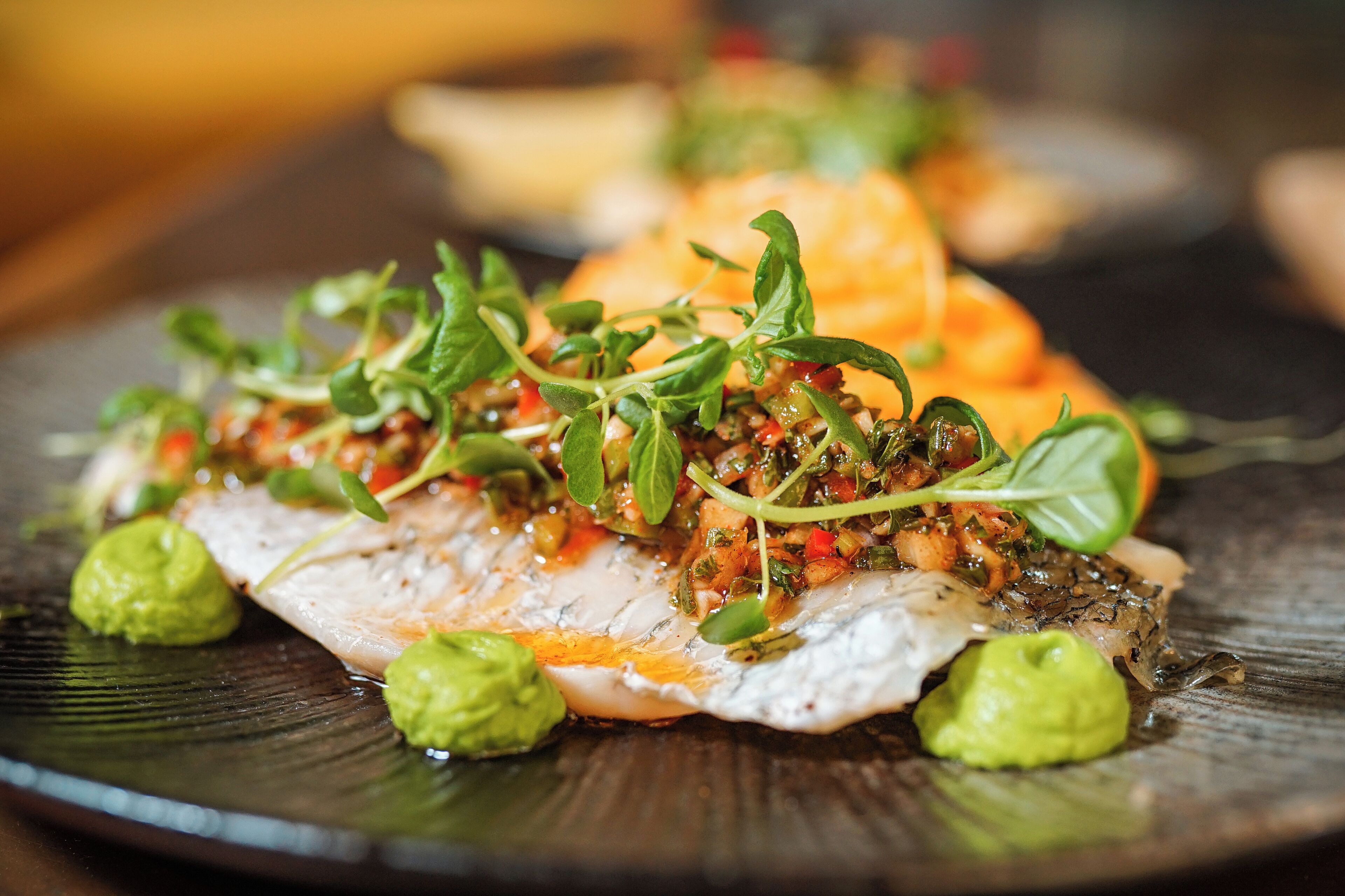 Un plato de comida con un trozo de pescado cubierto de vegetales verdes y salsa, decorado con rebanadas de aguacate.