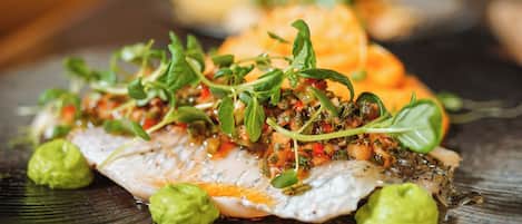 Un plato de comida con un trozo de pescado cubierto de vegetales verdes y salsa, decorado con rebanadas de aguacate.