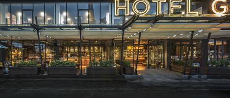 A hotel entrance with a large illuminated sign, glass doors, and outdoor seating areas with planters.