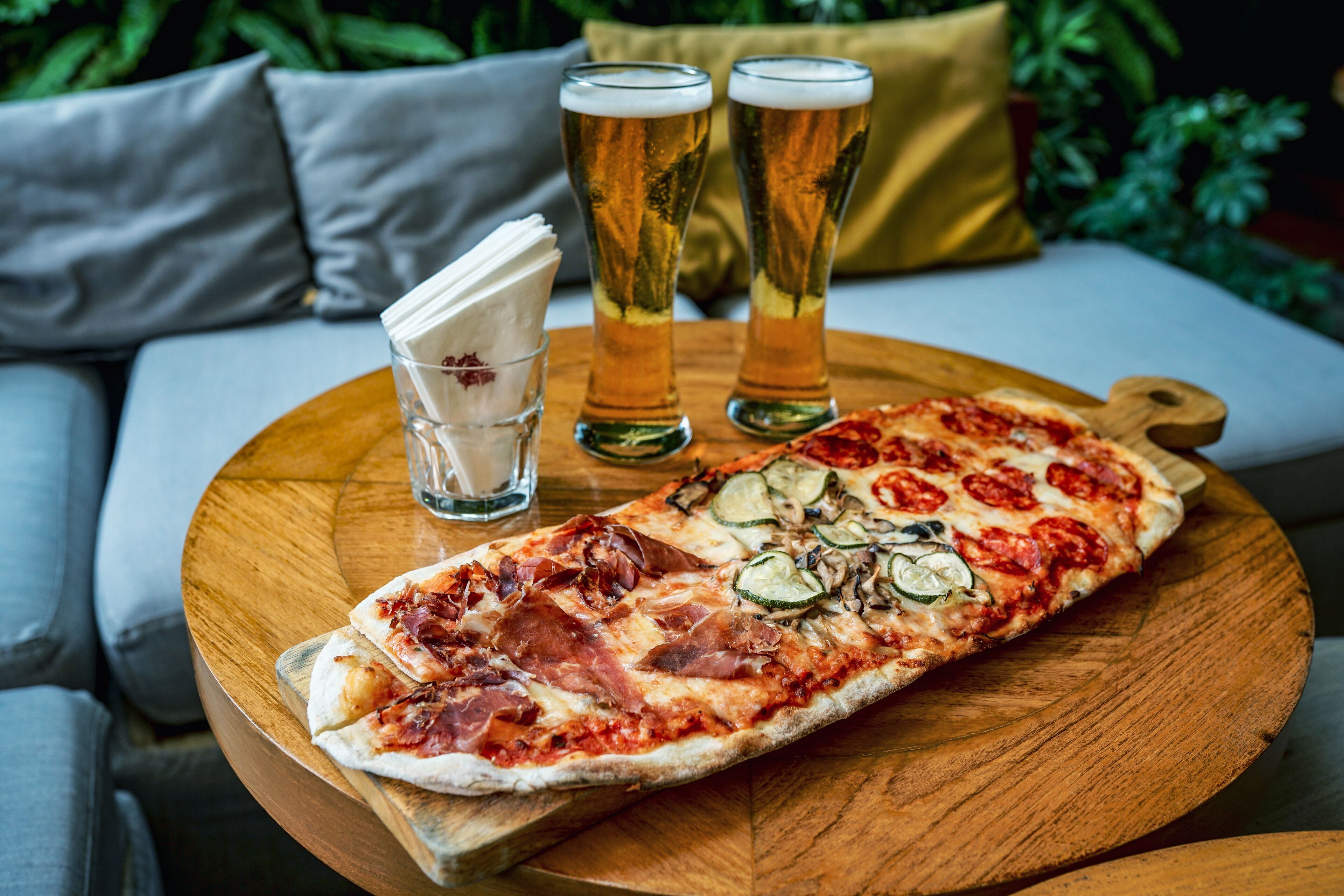 Una pizza grande sobre una tabla de madera, con una servilleta y dos vasos de cerveza en una mesa.