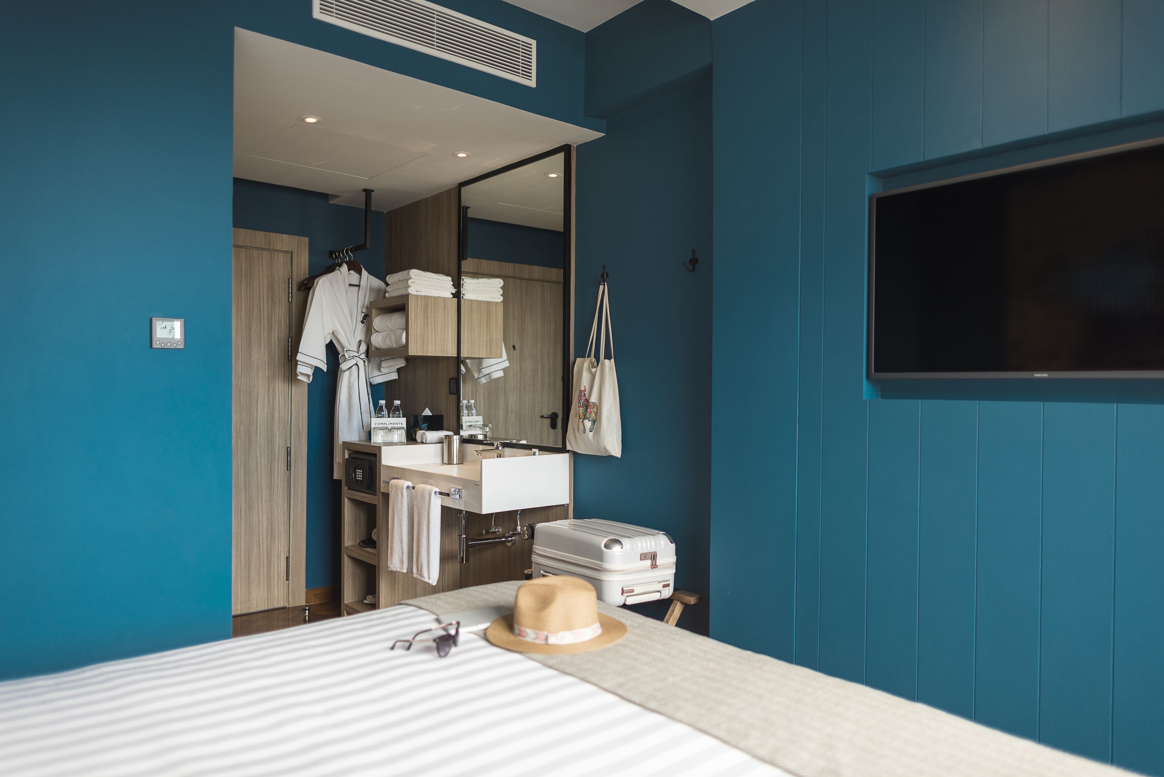 A hotel room with a bed, a suitcase, a hat, a mirror, a TV, and a bathroom visible through an open door.