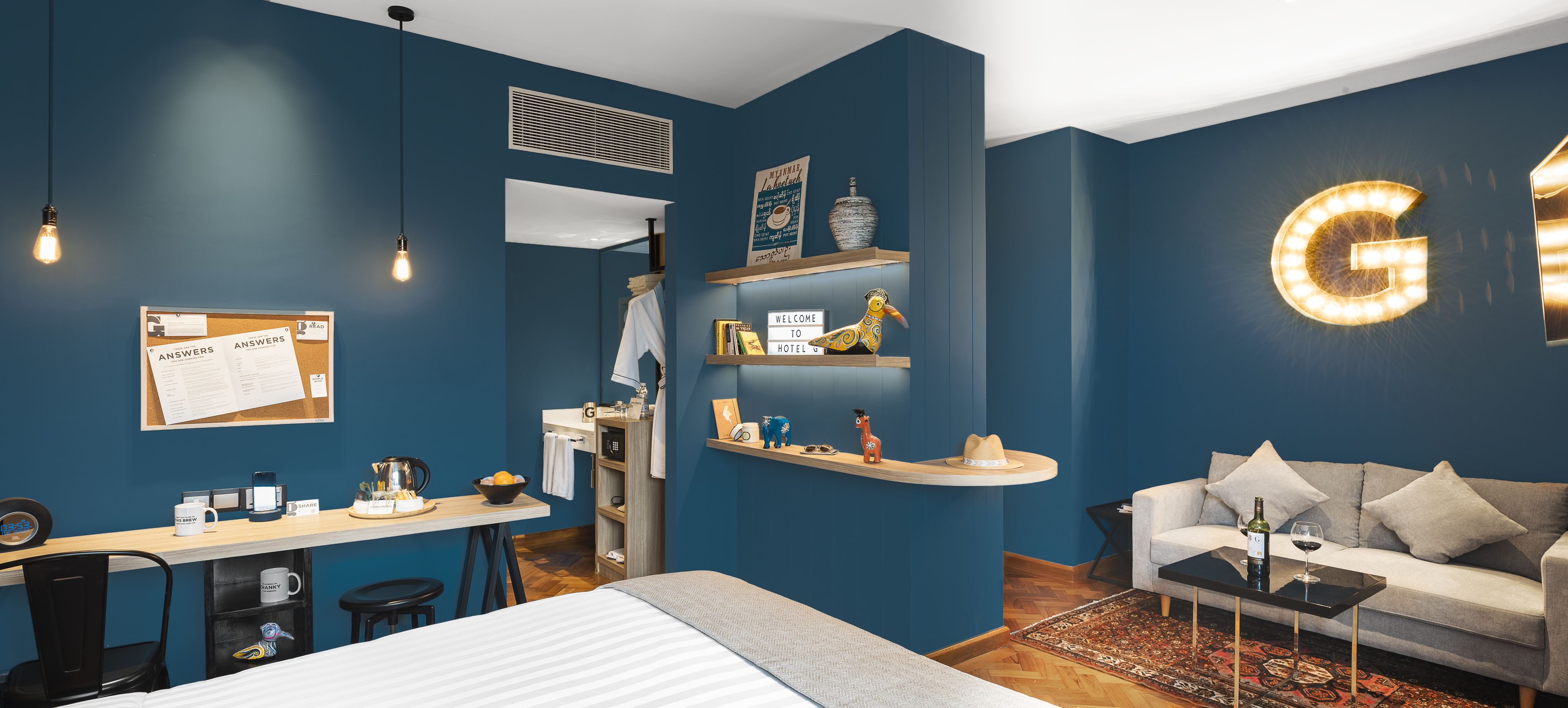 A modern hotel room with a blue wall, a bed, a desk with a chair, a sofa, and a coffee table. There is a bulletin board with a notice, a shelf with decorative items, and a large illuminated 'G' on the wall.