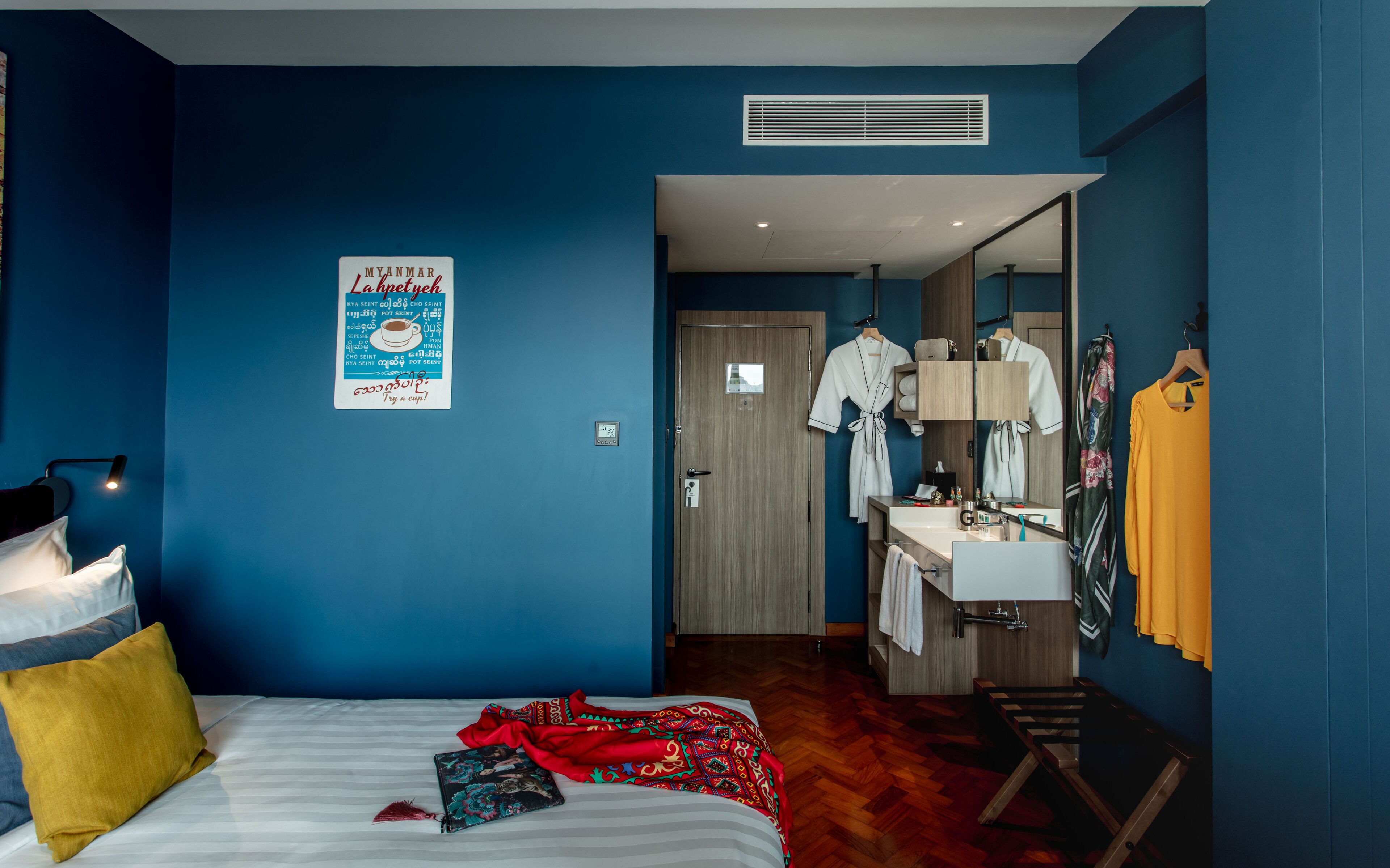 A hotel room with a bed, a bedside table, a mirror, a bathroom with a sink and a shower, and a poster on the wall.