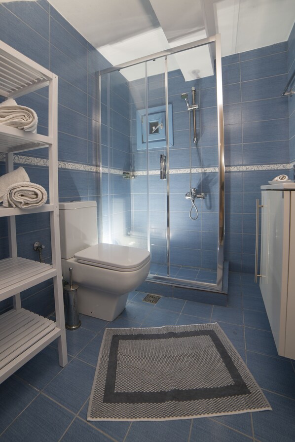 A bathroom with a glass-enclosed shower, a toilet, a sink, and a shelf with towels. - Almyra Guesthouses (Skiros)