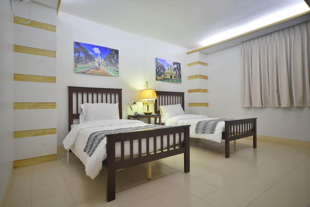 Ein Hotelzimmer mit zwei hölzernen Betten, weißer Bettwäsche, einem Nachttisch mit Lampe und zwei an der Wand hängenden Bildern.