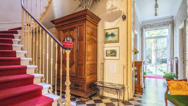 Un grand escalier recouvert d’une moquette rouge, un meuble en bois surmonté d’une vitre, et un sol à carreaux.