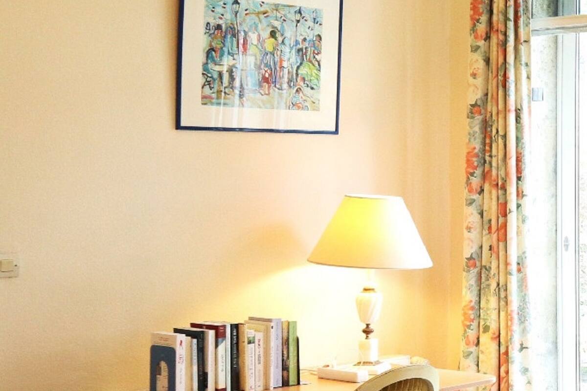 A study room with a desk, chair, books, a lamp, and a framed painting on the wall.