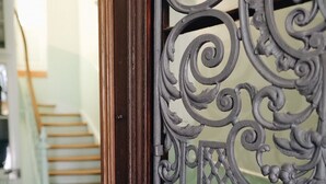 Ein Detailaufnahme eines verzierten Metalltors mit aufwendigen Mustern, das zu einer Treppe mit Holzboden und Schachbrettmuster führt.