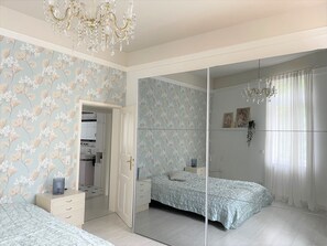 A bedroom with a mirrored closet, floral wallpaper, a chandelier, and a bed with a light blue bedspread. - Lovely Apartment in Baden-Baden (Baden-Baden)
