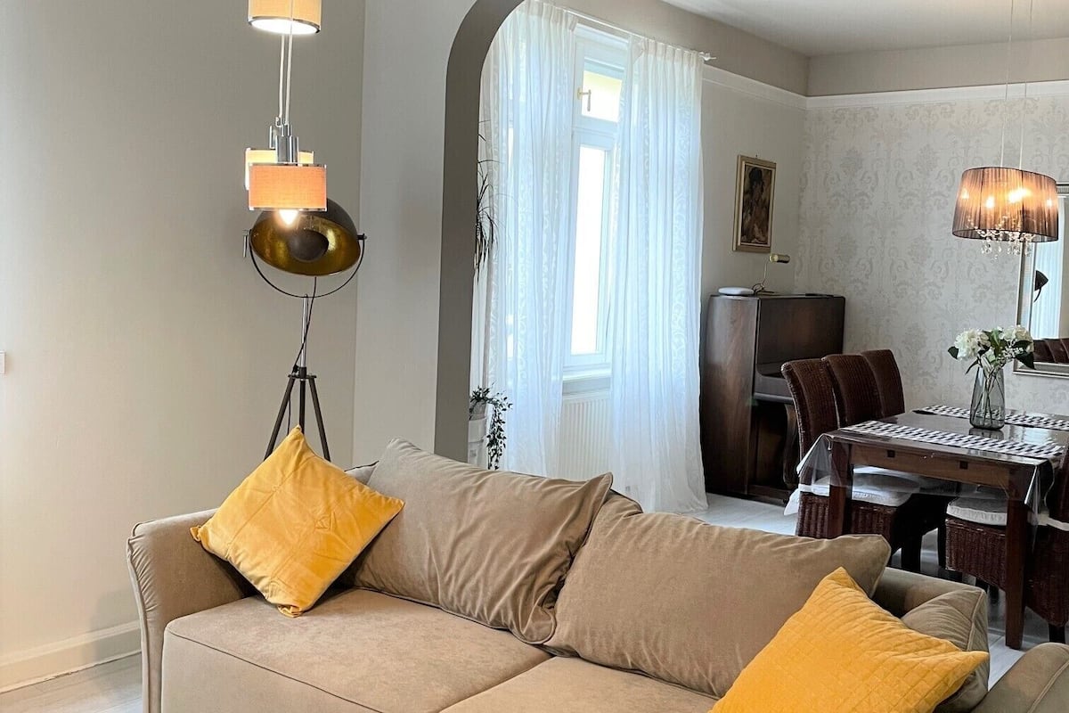A living room with a beige sofa, yellow cushions, a dining table with chairs, and a chandelier.