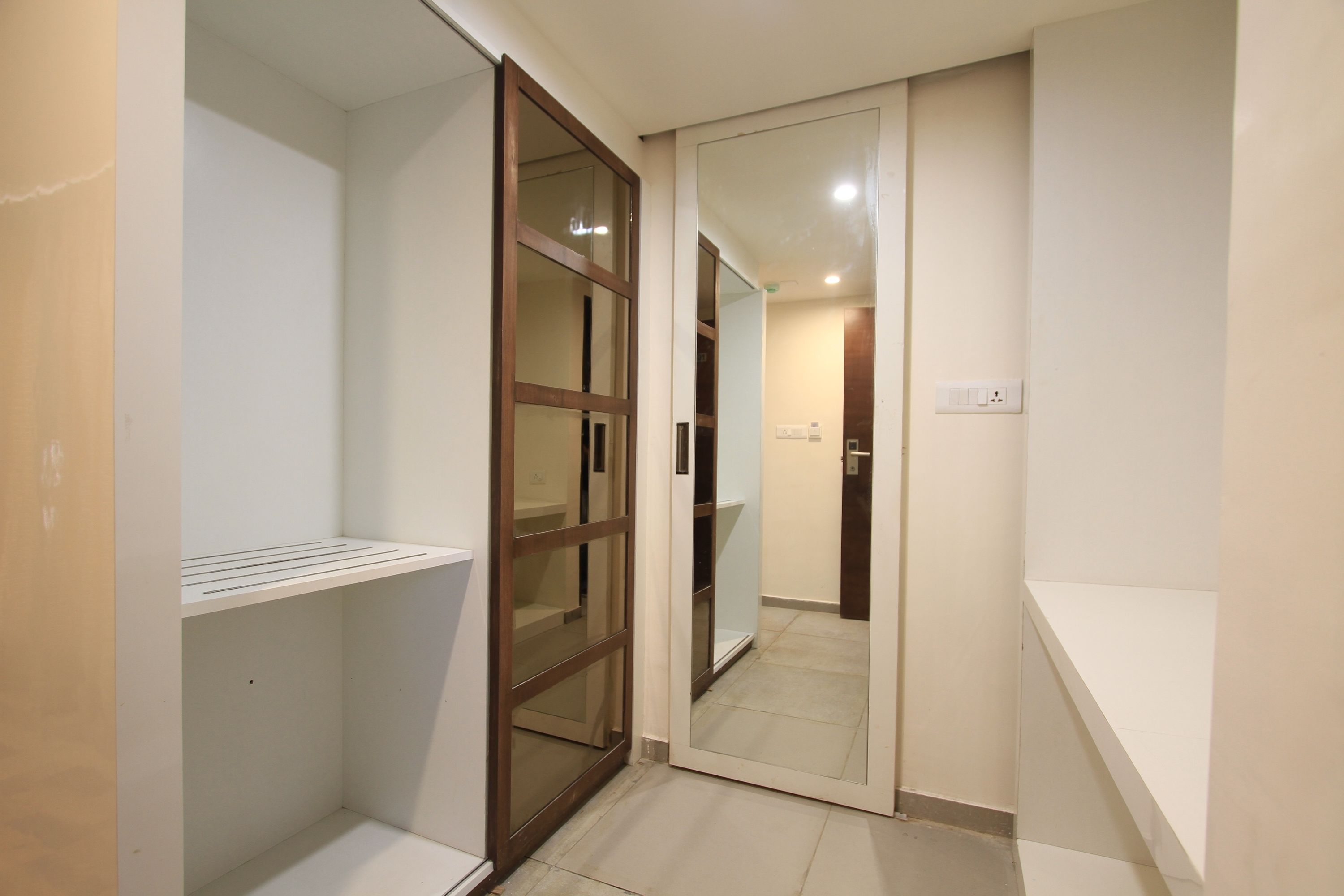 A narrow hallway with a sliding glass door leading to a room. There is a white shelf on the left and a white wall on the right.